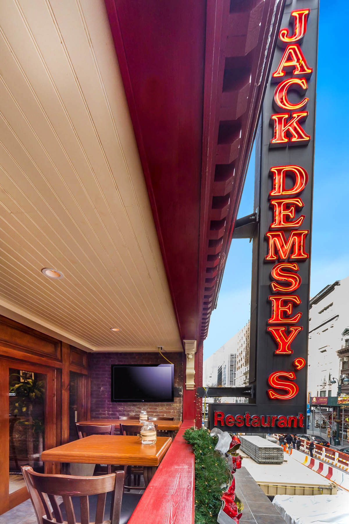 Outdoor seating area with wooden tables and chairs under a covered patio next to a neon Jack Demsey's Restaurant sign.