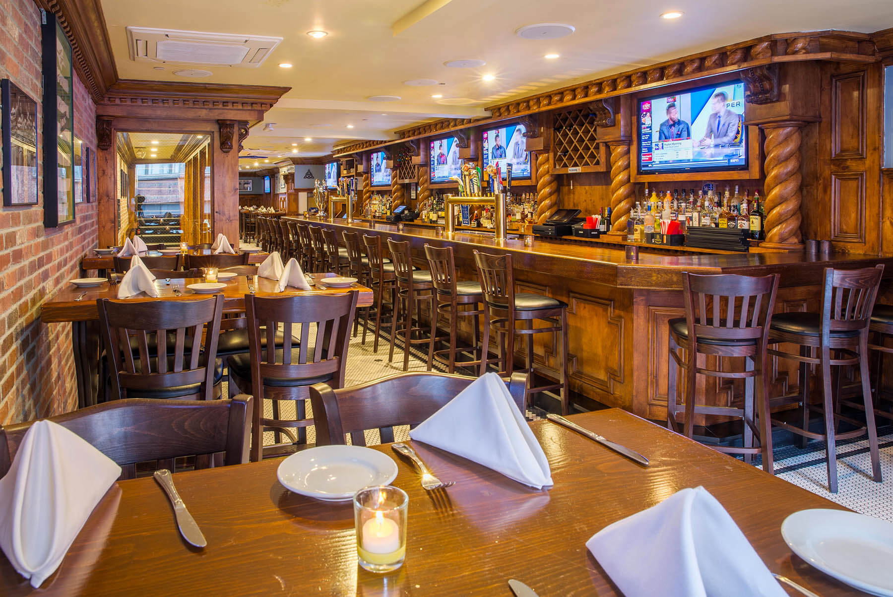 Cozy restaurant dining area with wooden tables set with white folded napkins, plates, cutlery, and a candle, adjacent to a wooden bar with high chairs and multiple TVs showing a sports program.
