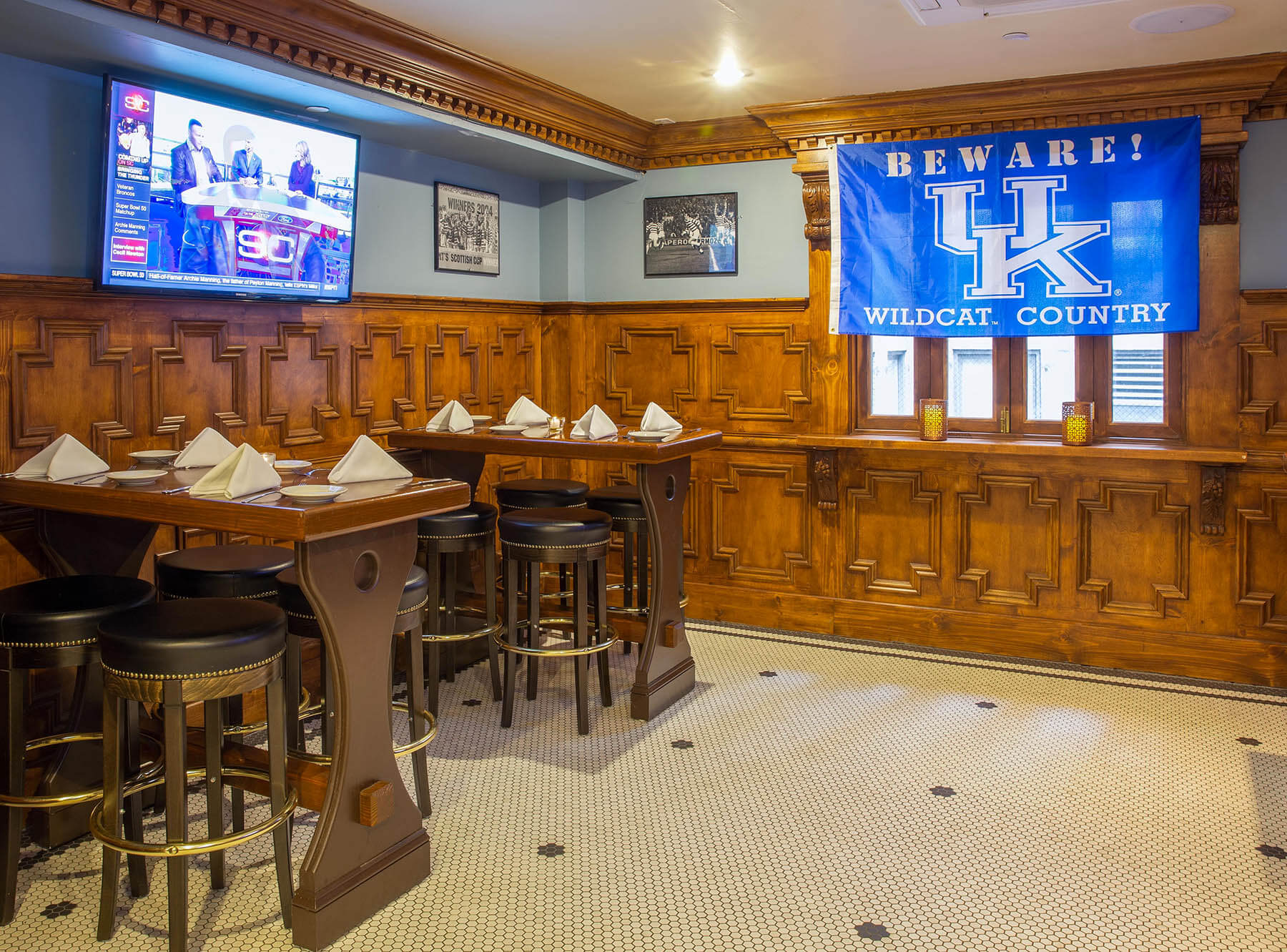 Corner dining room with wooden panel walls, a mounted TV, a blue UK Wildcats banner, and two set tables with stools.