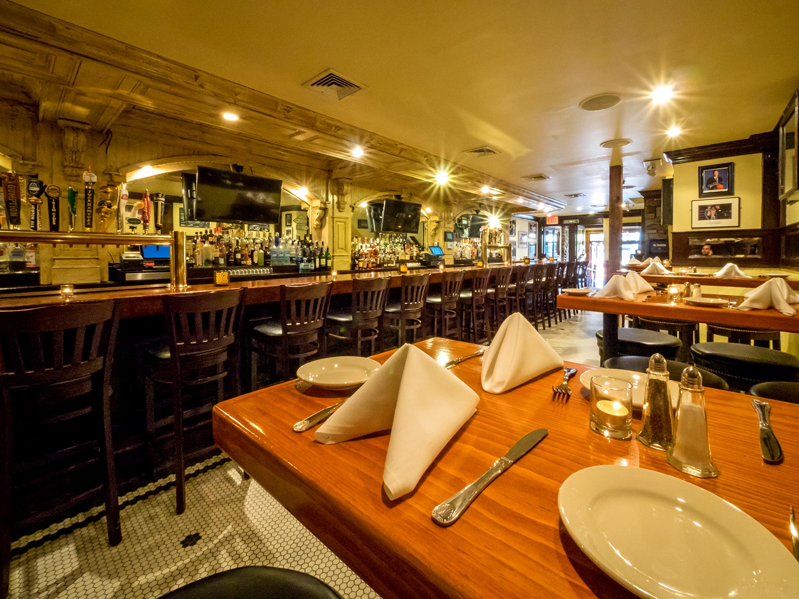 Warmly lit restaurant interior with a long wooden bar, bar stools, and neatly set tables with white napkins and utensils.