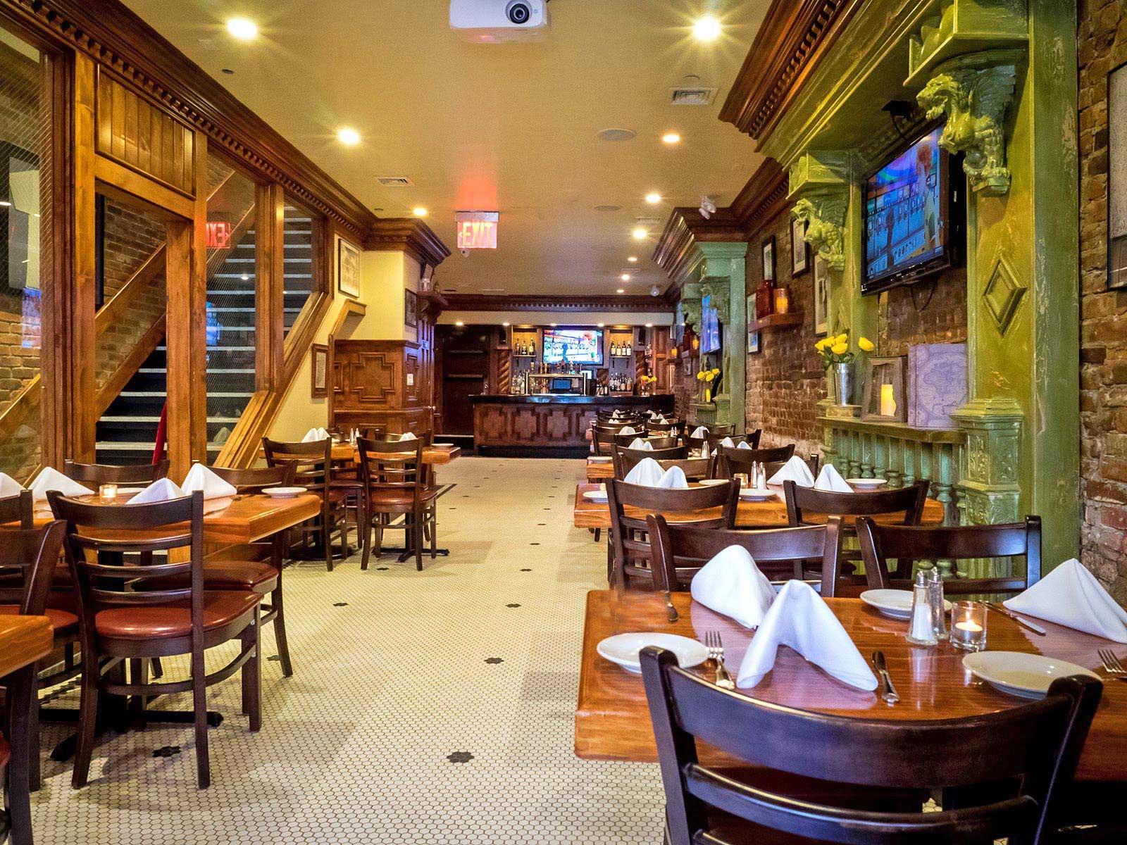 Cozy restaurant interior with wooden tables set with white napkins, dark chairs, exposed brick walls, and a bar in the background.